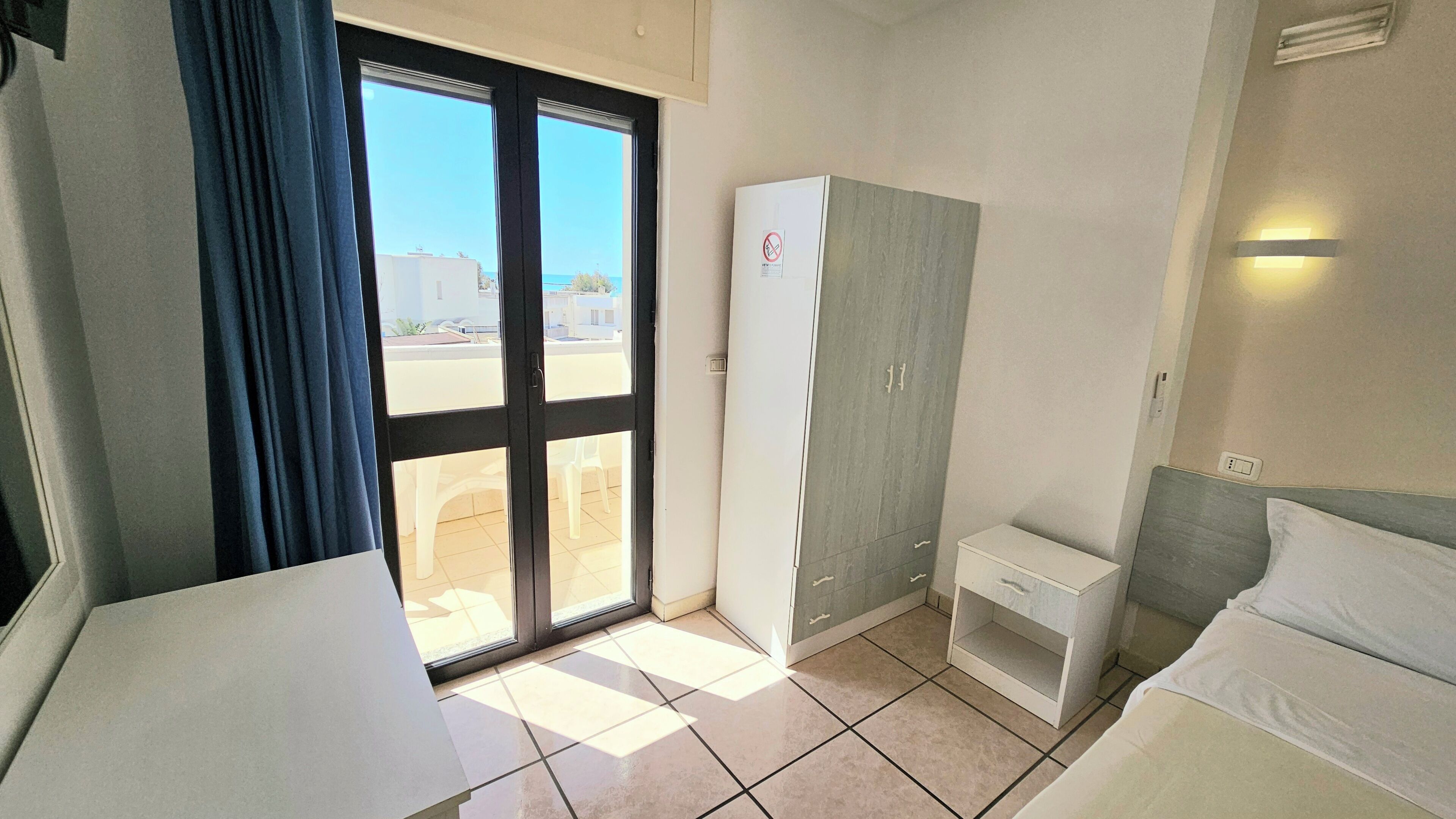 A hotel room with a bed, a wardrobe, a desk, and a window with a view of buildings and a clear sky.