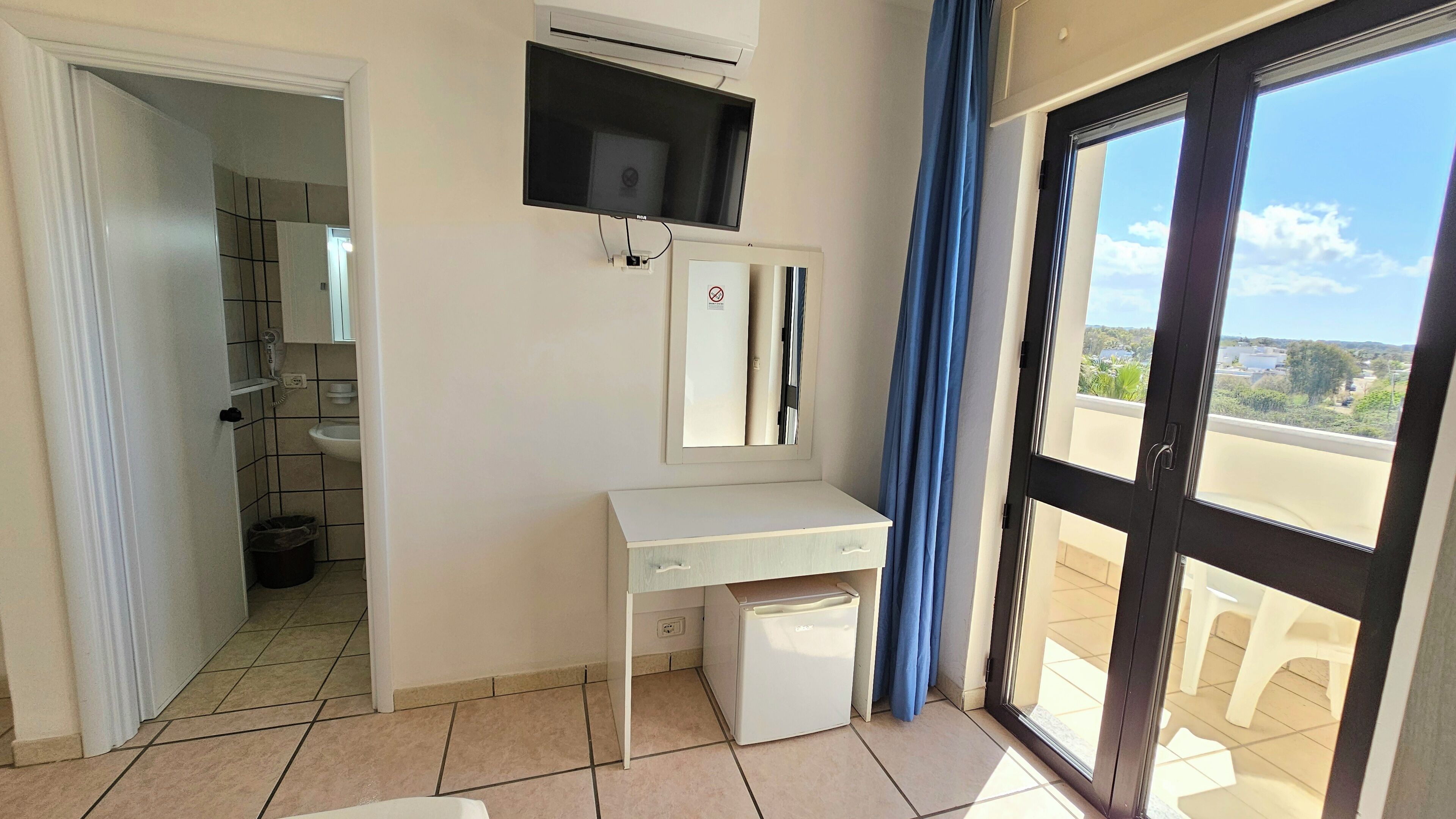 A room with a white desk, a mirror, a ceiling fan, a window with a view, and a door leading to a balcony.