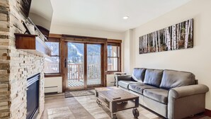 A spacious living room with a fireplace, a sofa, and a large window with a view of a snowy mountain. - Beautifully Updated 3 Bedroom in East Village - CS335 (Copper Mountain)