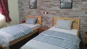 Una habitación con dos camas individuales, cada una con una colcha a rayas y almohadas a juego. Las camas tienen cabeceros de madera y están situadas frente a una pared de ladrillo. Dos cuadros enmarcados cuelgan de la pared sobre las camas.