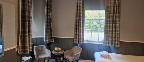 A hotel room with two single beds, a small table with a chair, and a window with curtains.