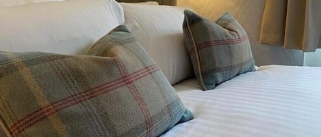 A hotel room with a large bed, multiple pillows, a headboard, a wall-mounted lamp, and a window with curtains.