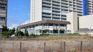 Ein Strandhotel mit mehreren Balkonen, ein sandiger Strand im Vordergrund und ein hohes Gebäude im Hintergrund.