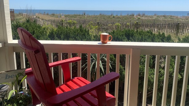 Ein roter Adirondack-Stuhl auf einem Balkon mit Blick auf das Meer und einem Holzzaun im Hintergrund.