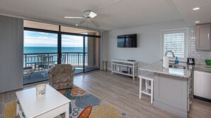 Una sala de estar moderna con un ventanal grande con vista al mar, un ventilador de techo, un televisor y una zona de cocina con un lavabo y encimera.