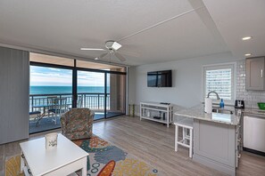 Un salon moderne doté d’une grande fenêtre donnant sur la mer, d’un ventilateur de plafond, d’un téléviseur et d’un coin cuisine avec un évier et un plan de travail.