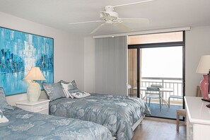 A bedroom with two beds, a ceiling fan, a painting, a nightstand, a desk, and a balcony with chairs. - Luxury, newly renovated 2 bedroom condo with a beautiful oceanfront balcony! (Ocean City)