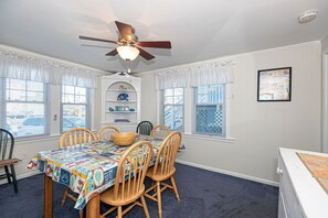Une salle à manger avec une table dressée pour six personnes, un ventilateur de plafond et une vue sur la rue grâce à de grandes fenêtres.