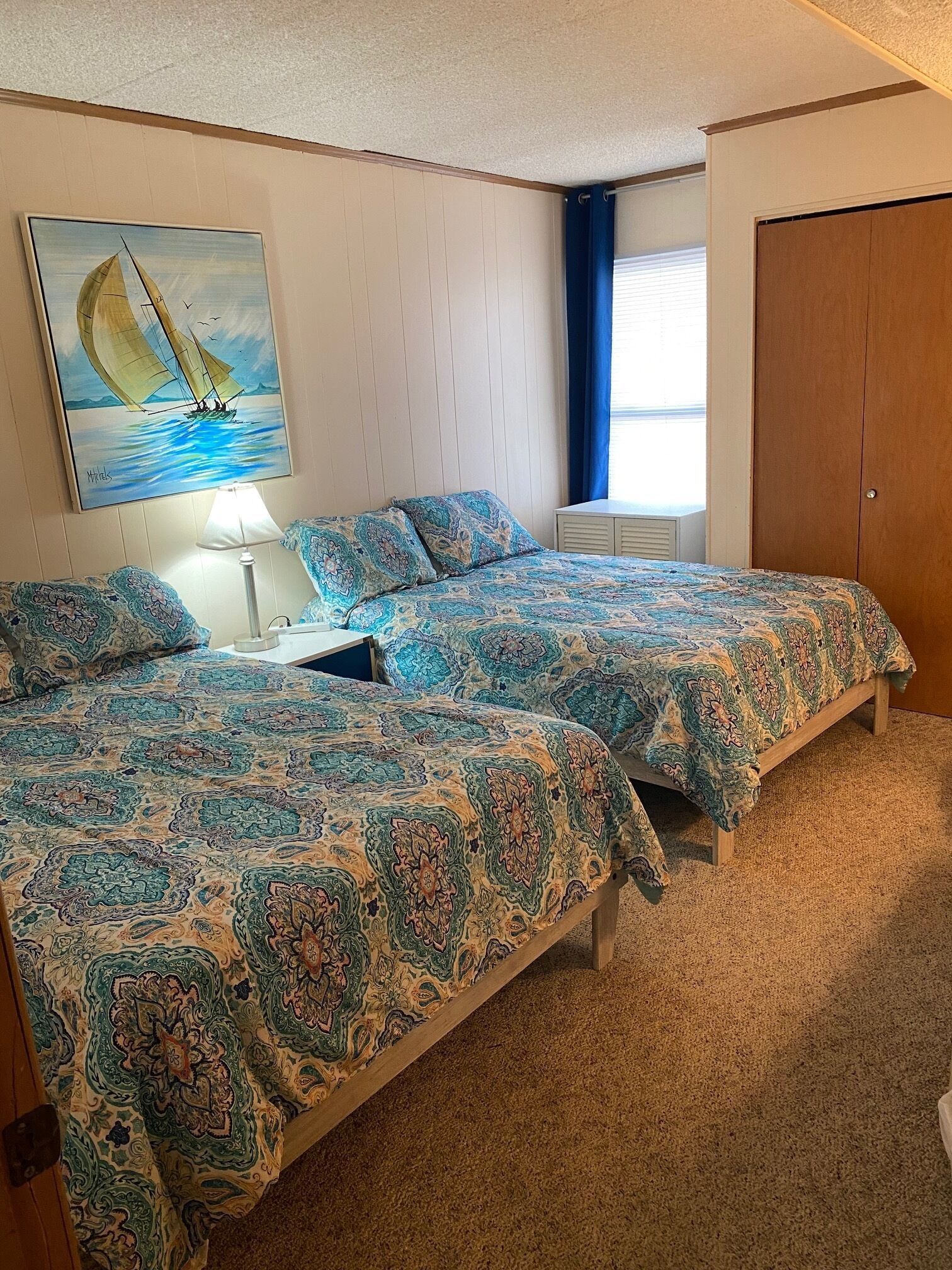 Une chambre avec deux lits, un tableau représentant un voilier, une lampe et une fenêtre avec des rideaux.