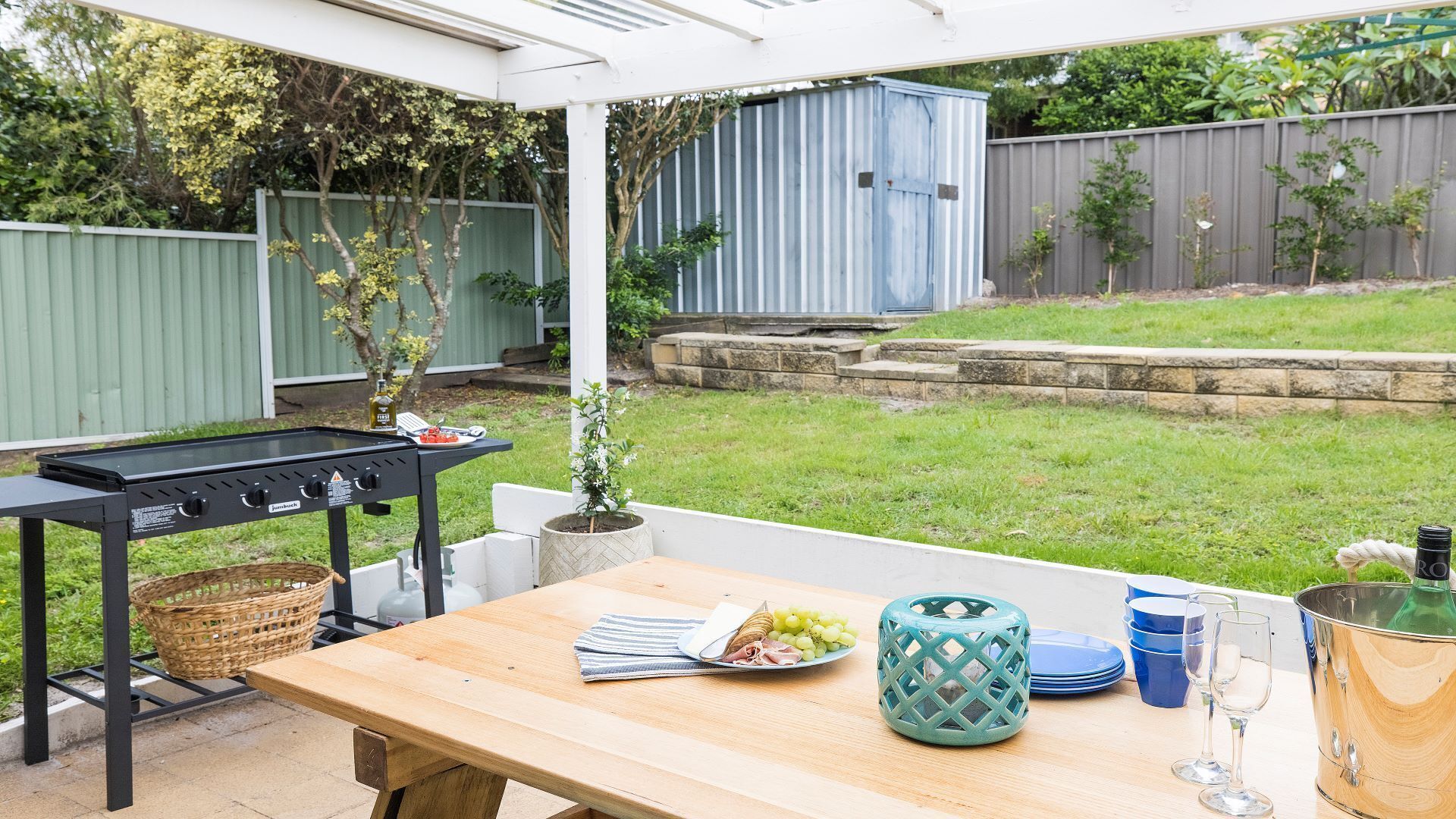 Eine Terrasse im Freien mit Grill, Tisch gedeckt mit Tellern und einer Flasche, und Blick auf den Garten.