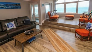 A spacious living room with a leather sofa, wooden coffee table, and orange armchairs. A large window offers a view of the ocean. - Stonecroft (Helston)