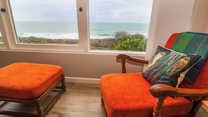 A room with a view of the ocean through large windows, a wooden rocking chair with a colorful cushion, and a matching ottoman. - Stonecroft (Helston)