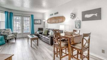 A living room with a sofa, a coffee table, a dining table with chairs, a lamp, and a wall with a fish decoration and a sign that says "BEACH HOUSE".