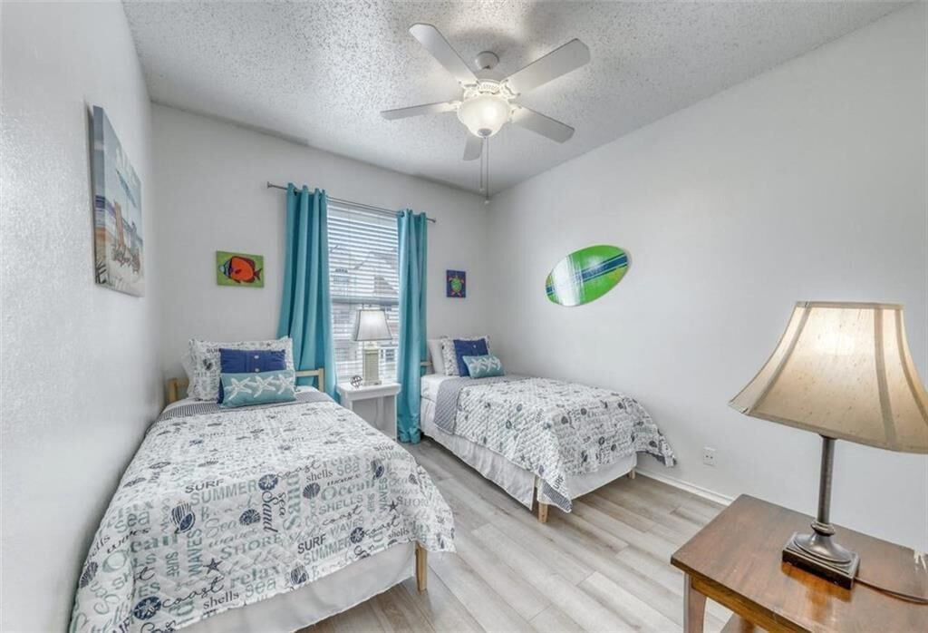 A bedroom with two beds, a ceiling fan, a window with curtains, and a lamp on a side table.