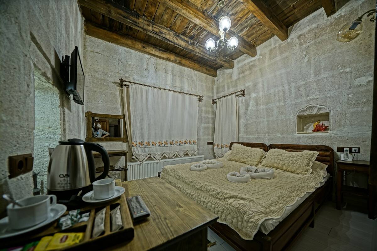 A cozy bedroom with a wooden bed, a bedside table with a kettle, cups, and a tray, and a wooden ceiling with exposed beams.