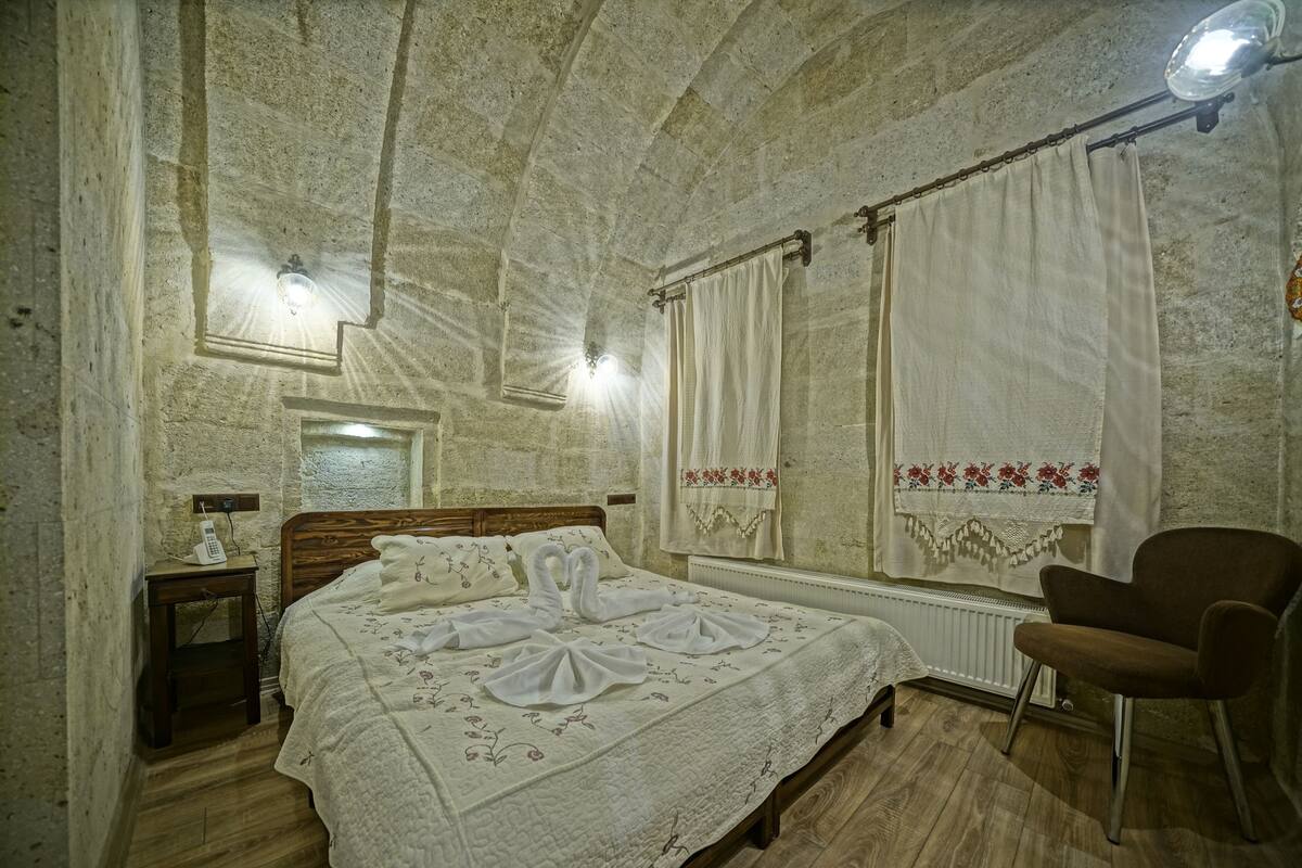 A room with a stone wall, a wooden bed, a chair, a nightstand with a phone, and a window with curtains.