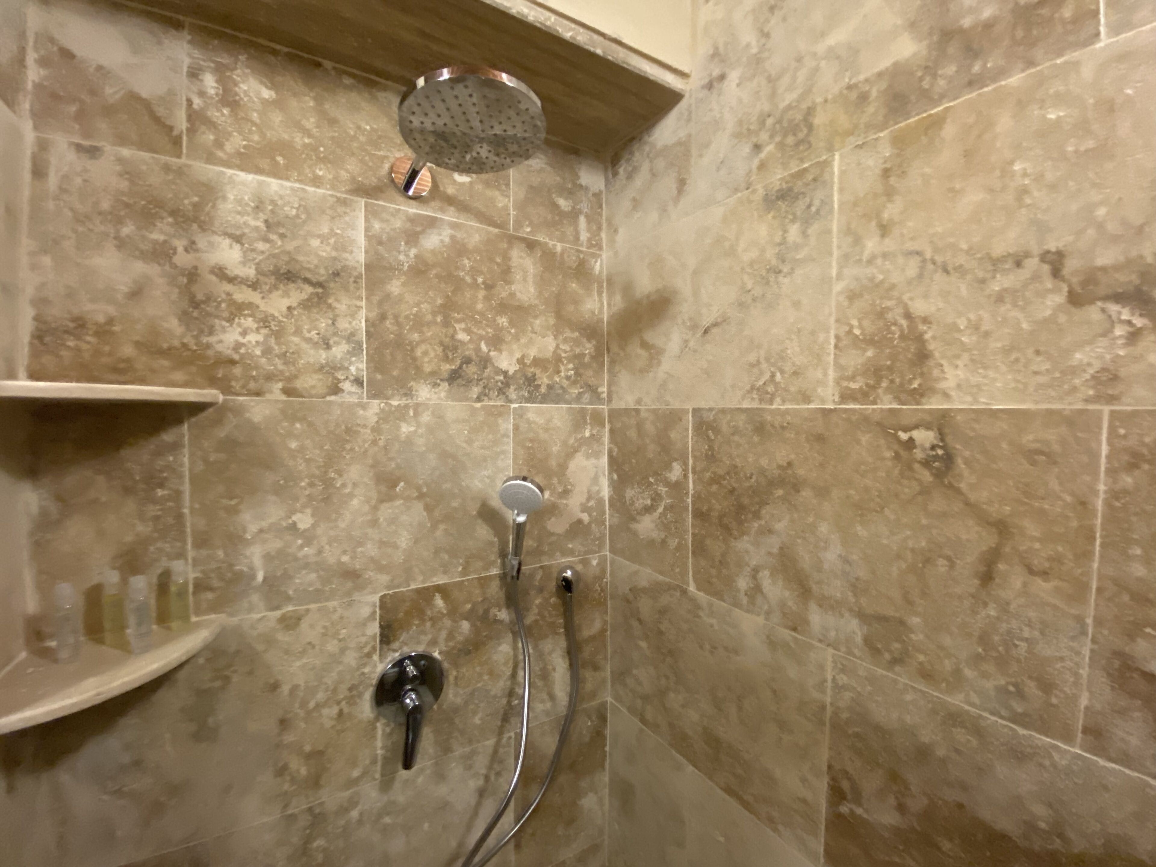 A bathroom with beige marble tiles, a showerhead, a handheld shower, and a shelf with toiletries.