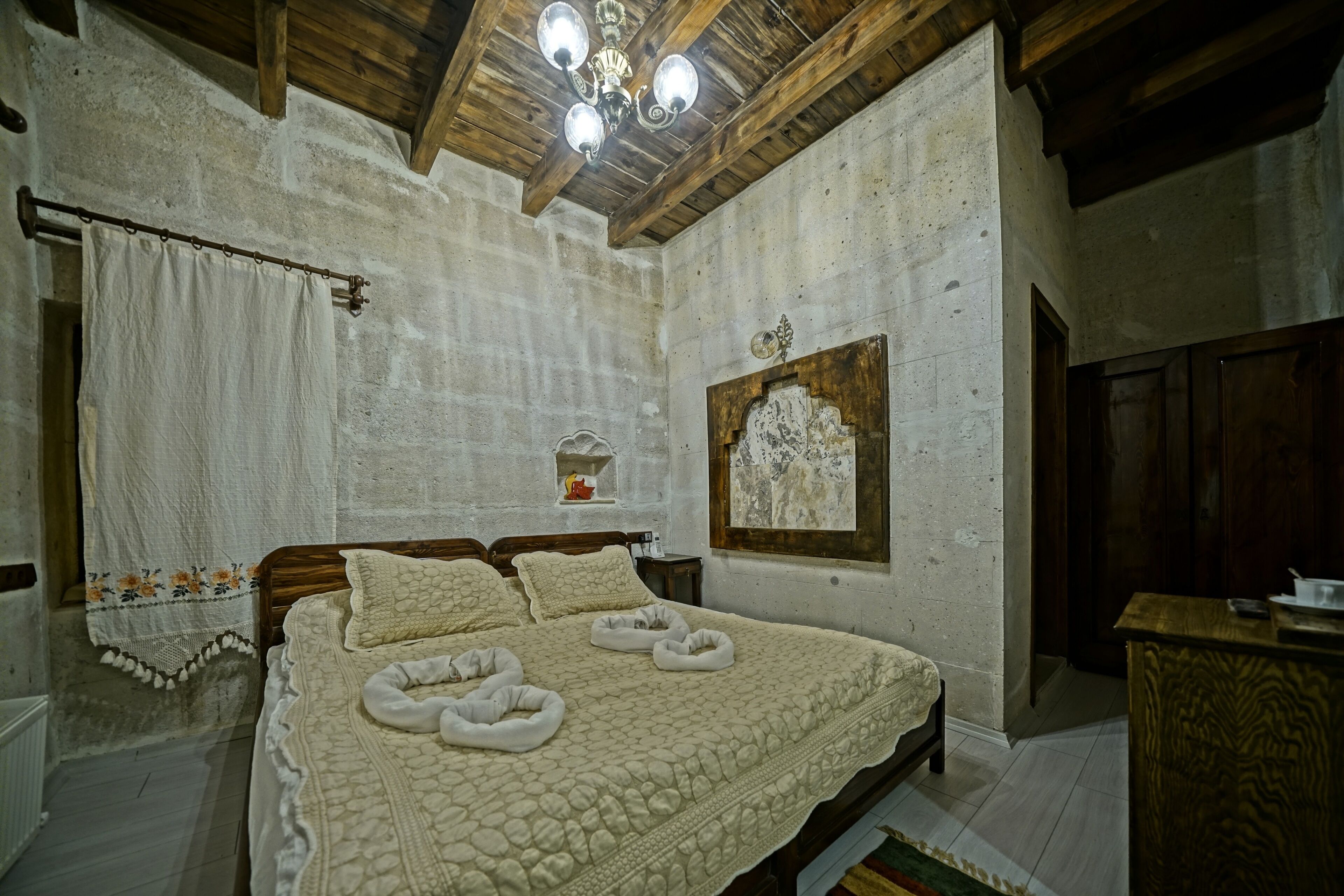 A bedroom with a large bed, wooden ceiling beams, a chandelier, and a stone wall.