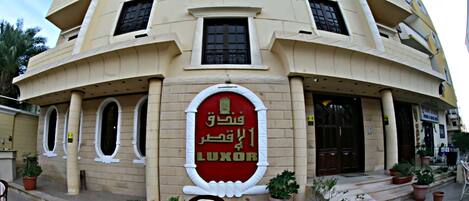 A building with a large sign reading "LUXOR" in Arabic, surrounded by potted plants and outdoor seating.