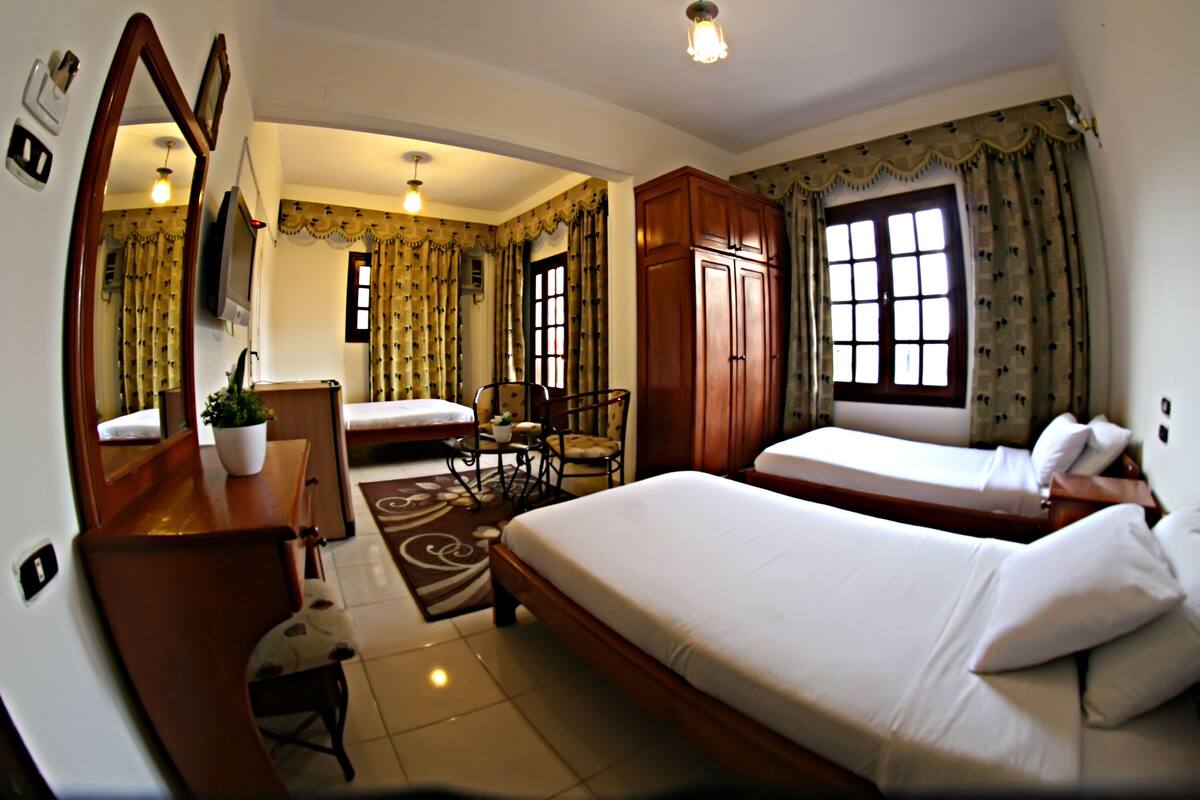 A hotel room with two beds, a wooden wardrobe, a desk with a chair, a mirror, and a television.