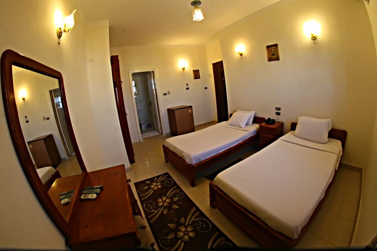 A hotel room with two single beds, a wooden desk with a chair, a large mirror, and a door leading to a bathroom.