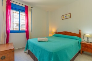 Een slaapkamer met een bed, een houten nachtkastje, een lamp, een raam met gordijnen en een aan de muur hangend schilderij.