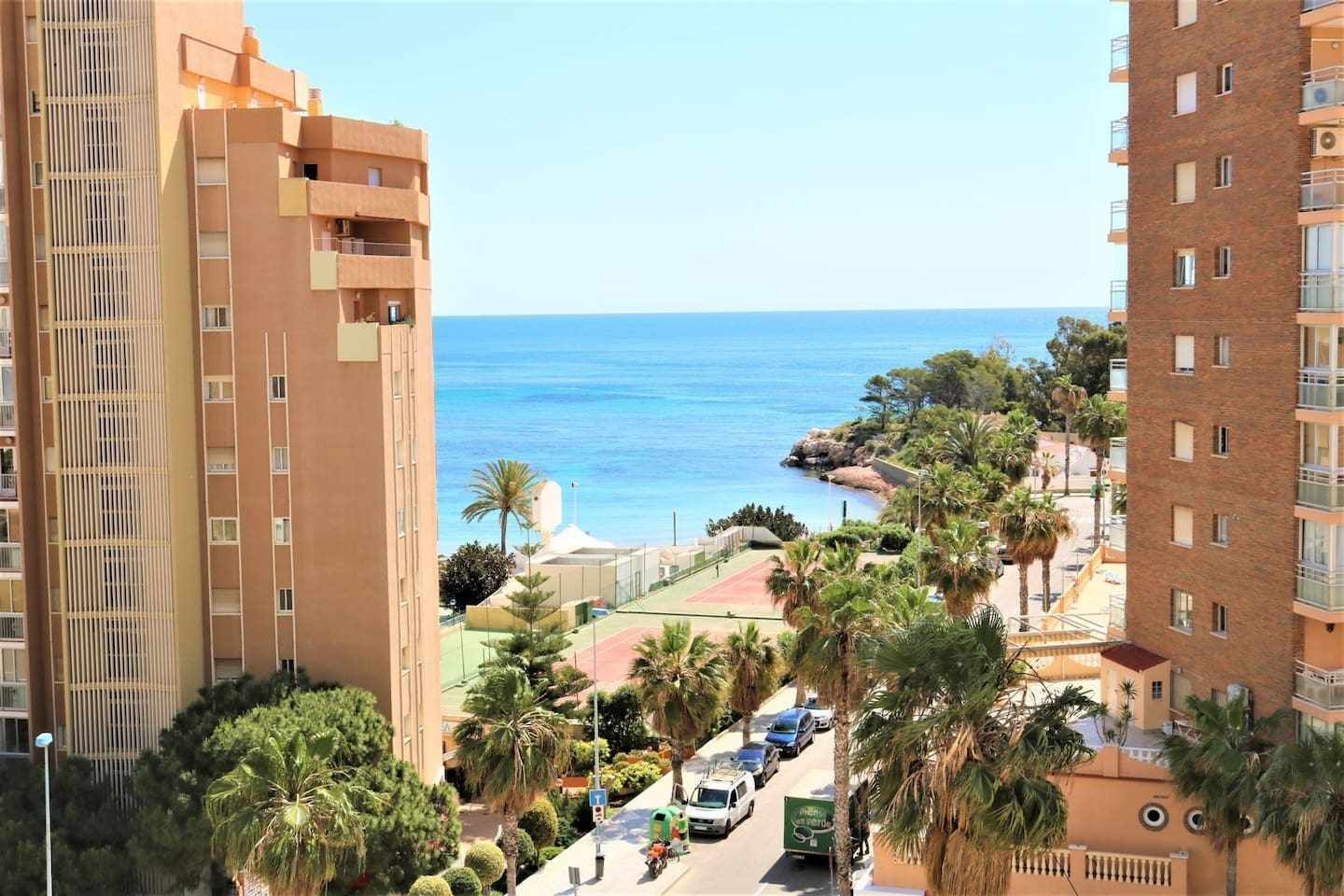 A coastal residential area with high-rise buildings, a clear view of the sea, and a road with parked cars.