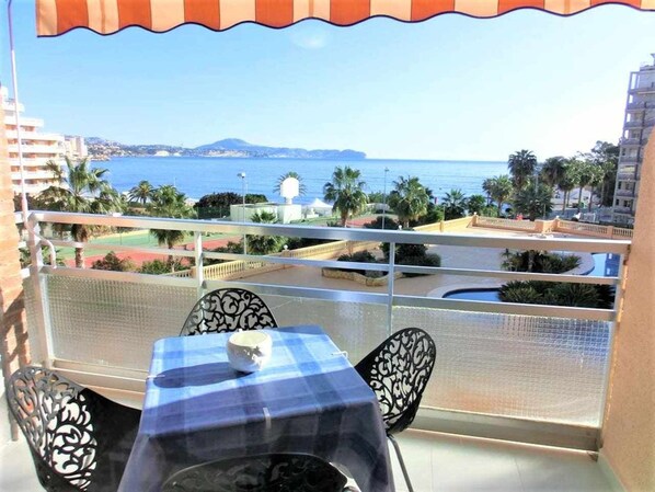 A balcony with a table and chairs, overlooking a pool and a view of the sea and distant hills. - Apartamento 1E 2 Dormitorios (Calpe/Calp)