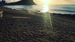Una spiaggia con una grande formazione rocciosa all'orizzonte, il sole che tramonta sull'oceano e un cielo sereno.