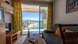 A modern living room with a sofa, a small table with a bottle and glasses, and a TV on a wooden cabinet. A large window offers a view of a beach and buildings. - mizzen 84, 3 bedrooms (Calpe/Calp)