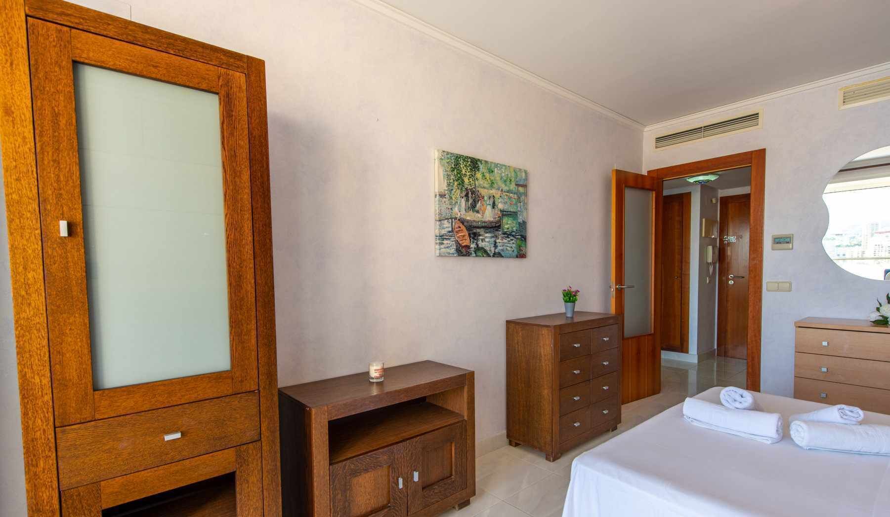 A hotel room with a large wooden wardrobe, a small wooden cabinet, a bed with white linens, a painting on the wall, and a mirror above the bed.
