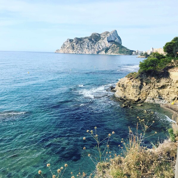 Un paysage côtier caractérisé par des eaux bleues et limpides, des falaises rocheuses et une montagne imposante qui se profile à l’horizon.