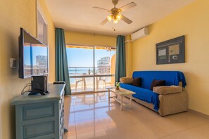 A room with a sofa, a TV, a ceiling fan, a balcony with a view of buildings, and a window with curtains.