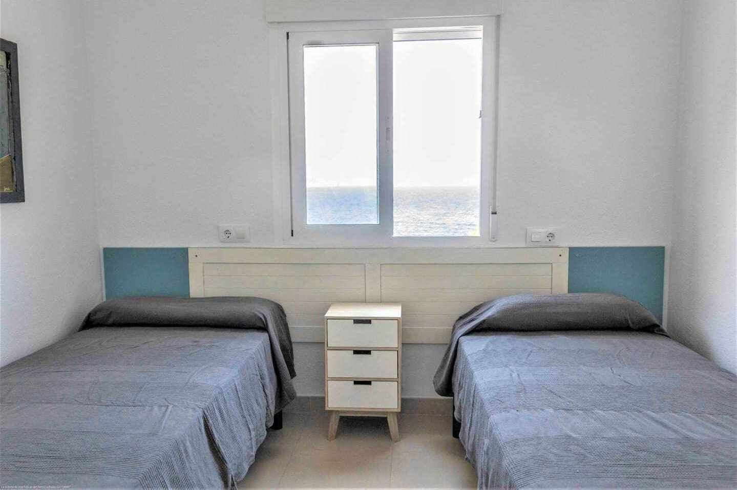 A bedroom with two beds, a window, and a small bedside table.