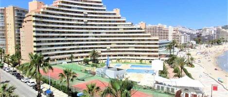 A multi-story hotel with a rooftop pool and tennis courts, surrounded by palm trees and a beach.