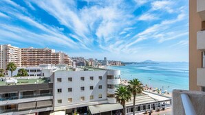 Paisagem costeira com vista para o mar, um edifício de vários andares com varandas, e uma área de praia com guarda-sóis.