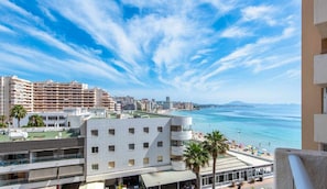 Un paisaje urbano costero con vista al mar, un edificio de varios pisos con balcones, y una zona de playa con sombrillas.