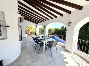 Un espace de restauration extérieur abrité, avec vue sur la piscine, comprenant une table dressée pour quatre personnes, entourée de chaises.