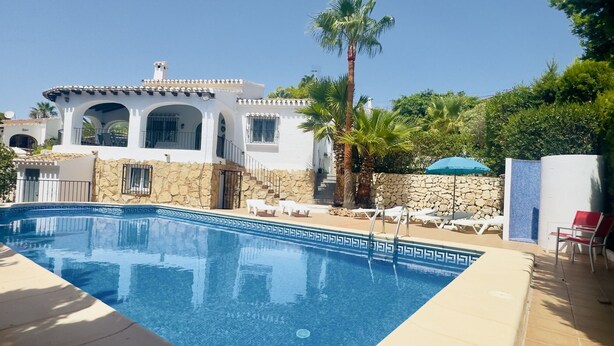 Una piscina con muro de piedra y palmeras, un edificio blanco con arcos, y un cielo azul despejado.
