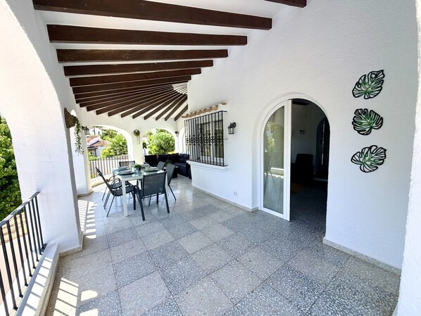 Ein überdachter Patio-Bereich mit Esstisch und Stühlen, einer Couch und einer Wand mit Blättermotiv.