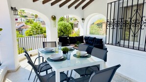 Un espacio comedor al aire libre cubierto con una mesa preparada para dos personas, sillas alrededor y un sofá con cojines.