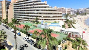 A multi-story hotel with a rooftop pool and tennis courts, surrounded by palm trees and a beach.