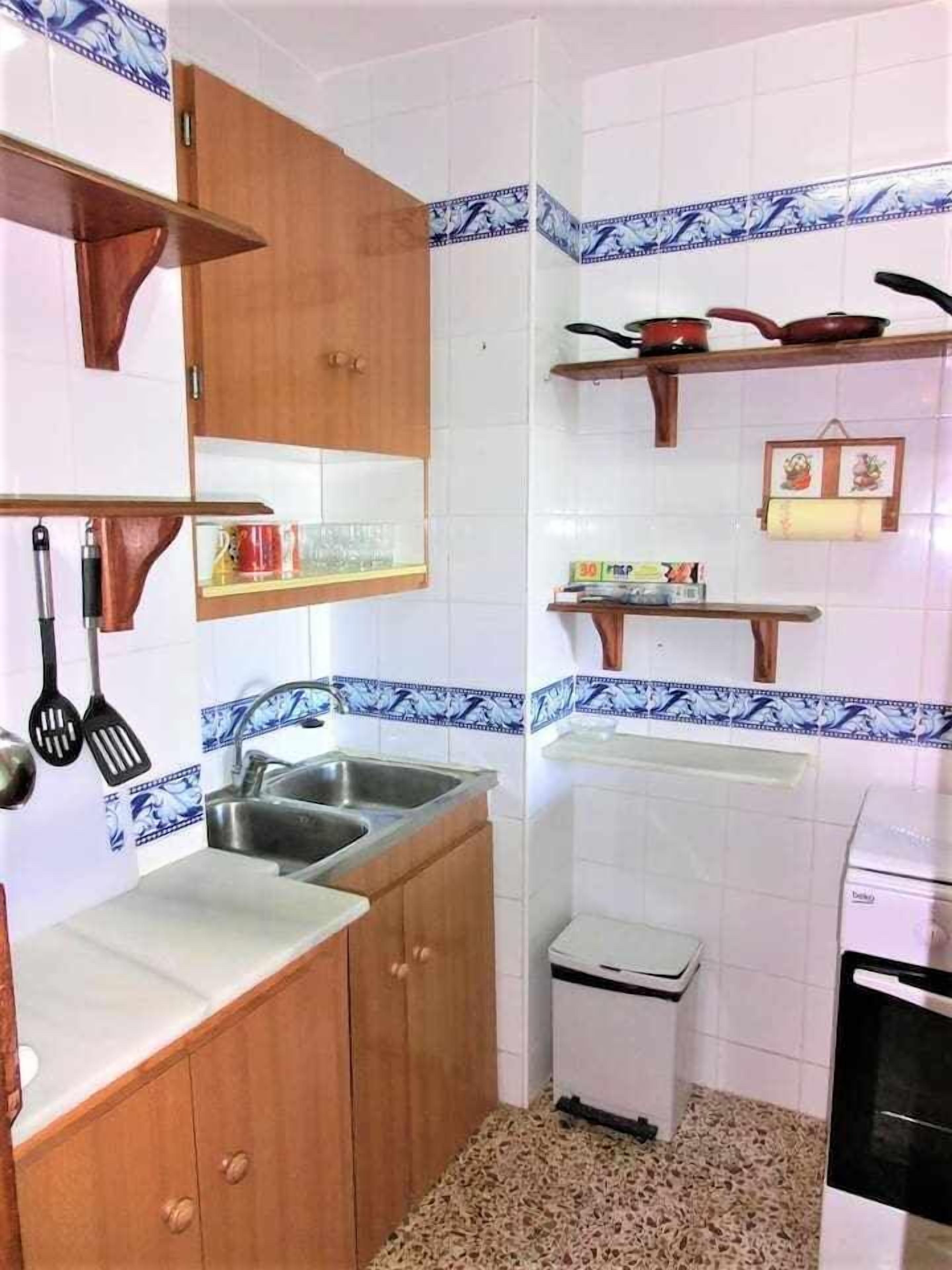 Cocina compacta con armarios de madera, un lavamanos, un pequeño cubo de basura y una estufa.