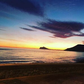 A sunset over a calm sea with distant mountains and a stone wall in the foreground. - 14B Vora 1st line of the beach with sea views (Calpe/Calp)