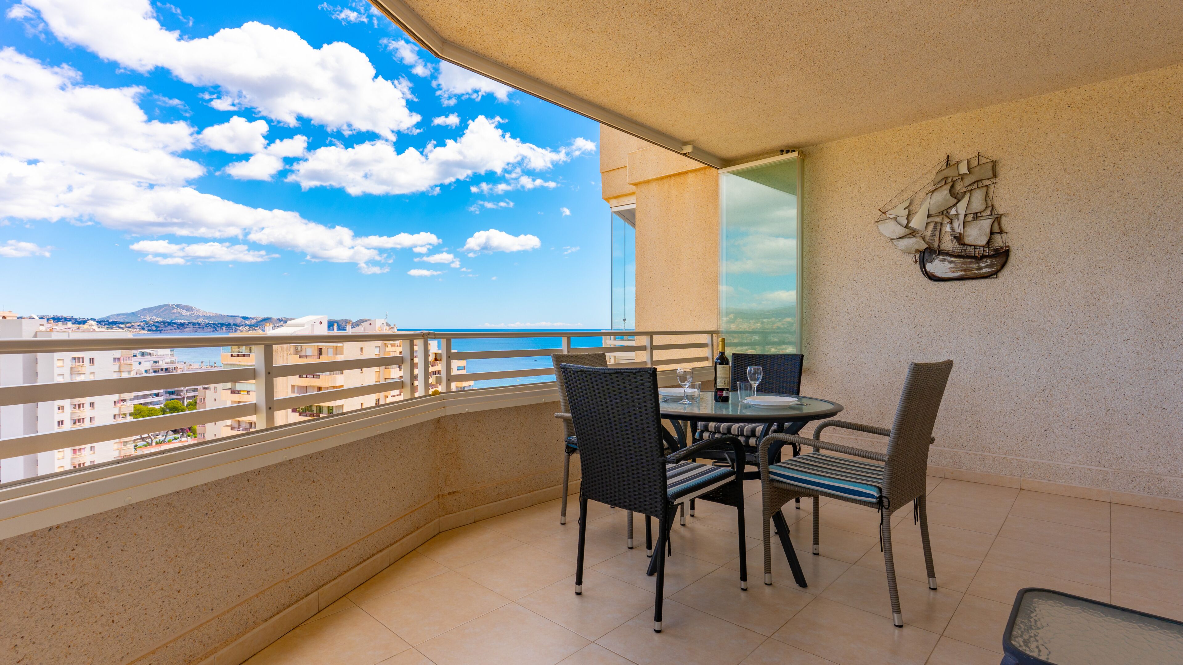 A balcony with a table and chairs, a ship decoration, and a view of the sea and buildings.