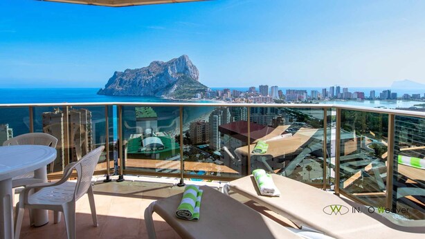 Un balcón con barandilla de cristal, una mesa, sillas y una tumbona. Se ve una montaña y el horizonte de la ciudad al fondo.