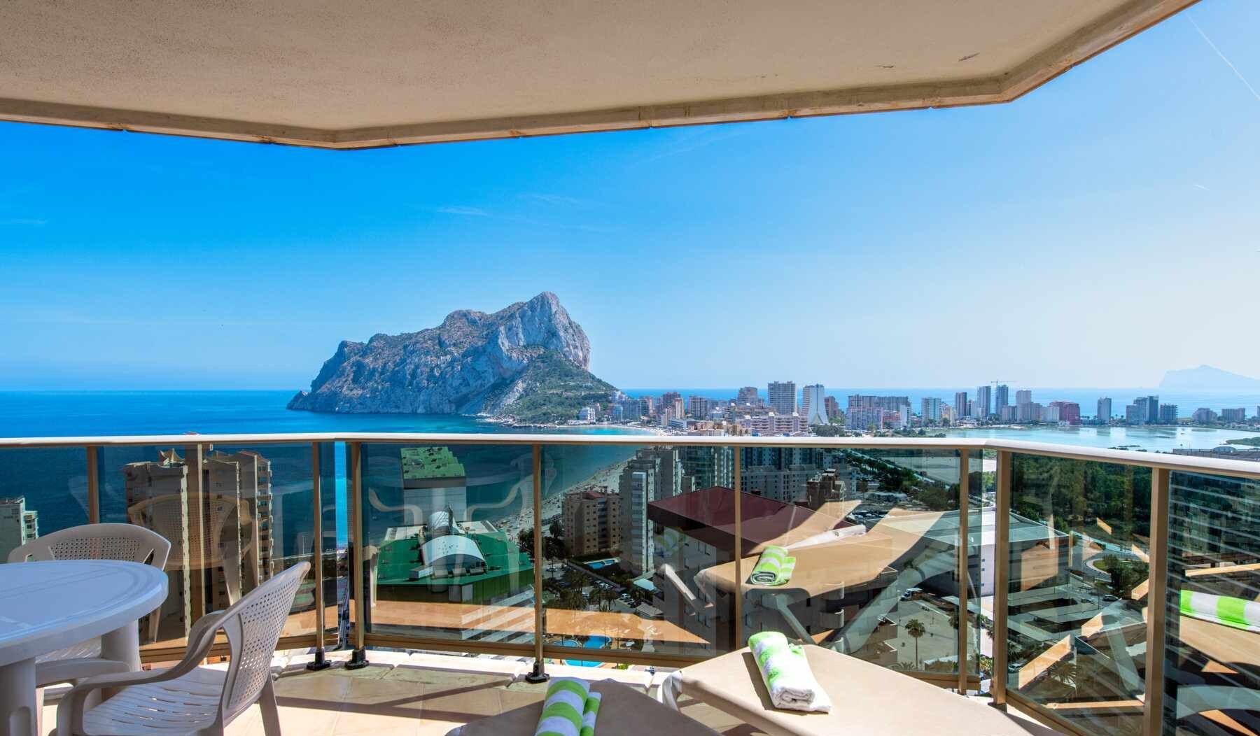 A balcony with a glass railing, a table, chairs, and a sun lounger. A mountain and city skyline are visible in the background.