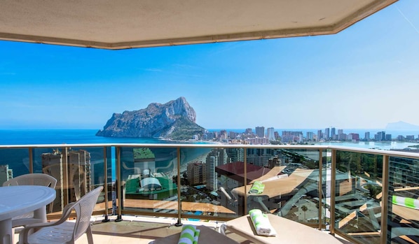 Un balcon doté d’une balustrade en verre, d’une table, de chaises et d’une chaise longue. On aperçoit en arrière-plan un paysage montagneux et la silhouette de la ville.