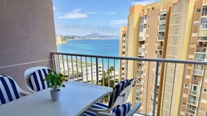 A balcony with a white table and striped chairs, overlooking a coastal cityscape with high-rise buildings.