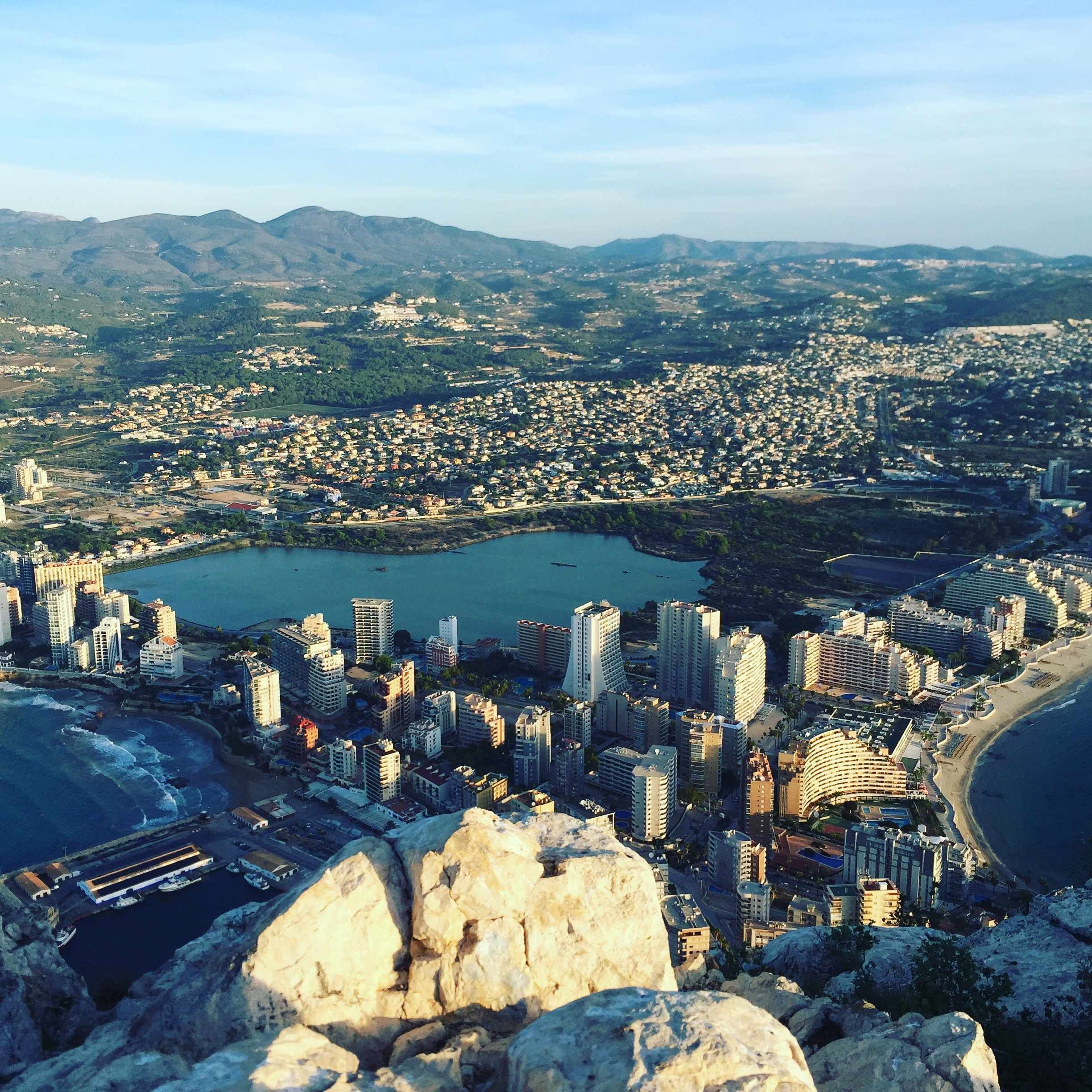 A coastal cityscape with high-rise buildings, a large body of water, and mountainous terrain in the background.
